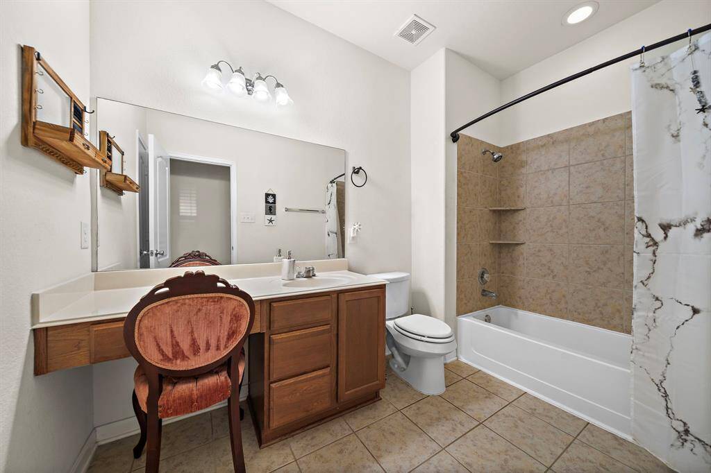 Modern bathroom with neutral tones featuring a large mirror, stylish lighting, and a combination shower-bathtub with tile surround. The space is accessorized with a unique chair and has ample counter space and storage.