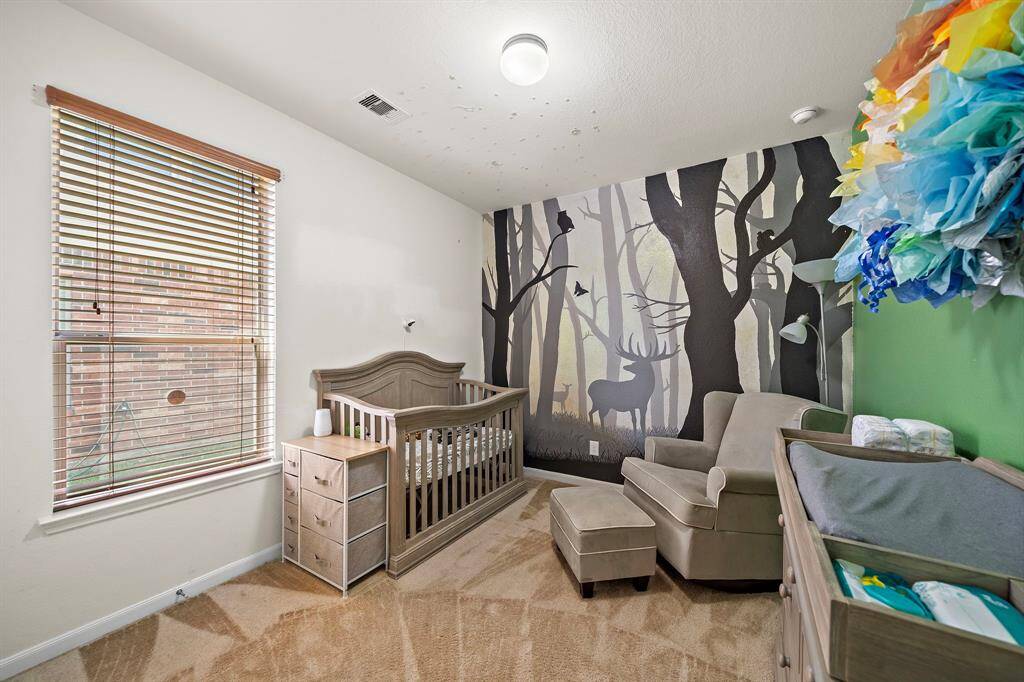 Cozy nursery with woodland-themed mural, featuring a crib, comfortable armchair, and changing table. The room is well-lit with a large window and has soft carpet flooring.