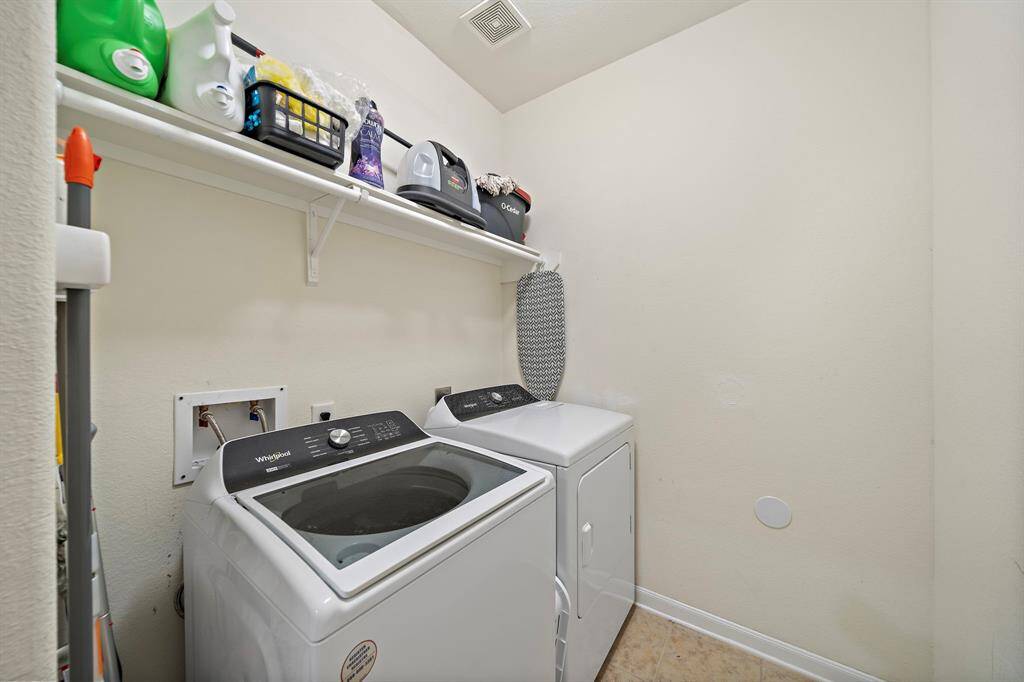 This photo shows a compact laundry room featuring a washer and dryer with a shelf above for storage. The space is well-organized, with cleaning supplies and an ironing board neatly arranged.