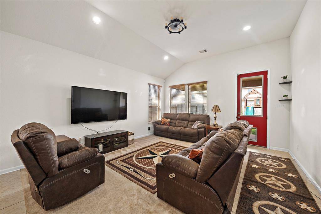This spacious living room features high ceilings, a wall-mounted TV, and comfortable seating. Large windows provide natural light and a view of the backyard. The decor includes a patterned rug and modern lighting, creating a cozy and inviting atmosphere.