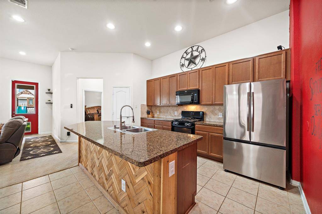 This kitchen features a spacious island with granite countertops, modern stainless steel appliances, and ample wooden cabinetry. The open layout connects to a cozy living area, and a vibrant red accent wall adds a touch of color.