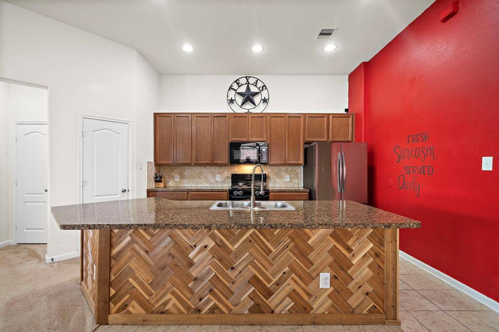 This kitchen features a spacious granite island with a unique herringbone wood design. It has modern appliances, warm wood cabinetry, and a bold red accent wall with playful decor. Ideal for cooking and entertaining.