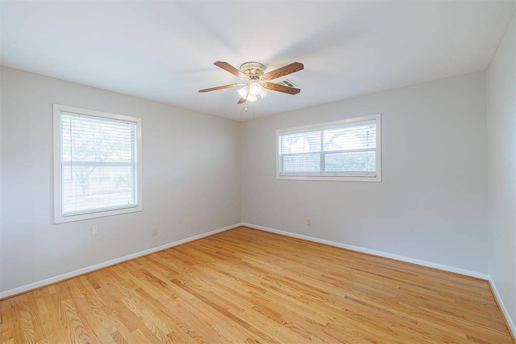 This bright, spacious bedroom features hardwood floors, neutral walls, and ample natural light from two windows. A ceiling fan adds comfort and style.