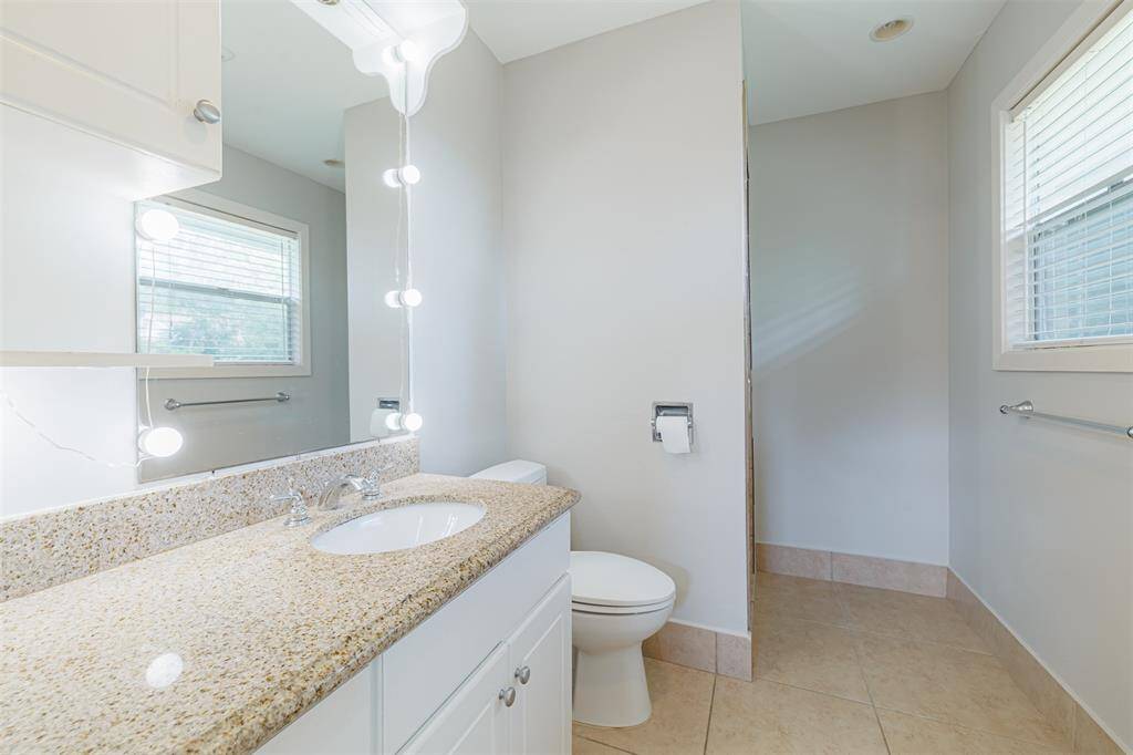 This primary bathroom features a granite countertop with a modern sink, ample cabinet space, and a large mirror with stylish lighting. It includes a walk-in shower and windows for natural light.
