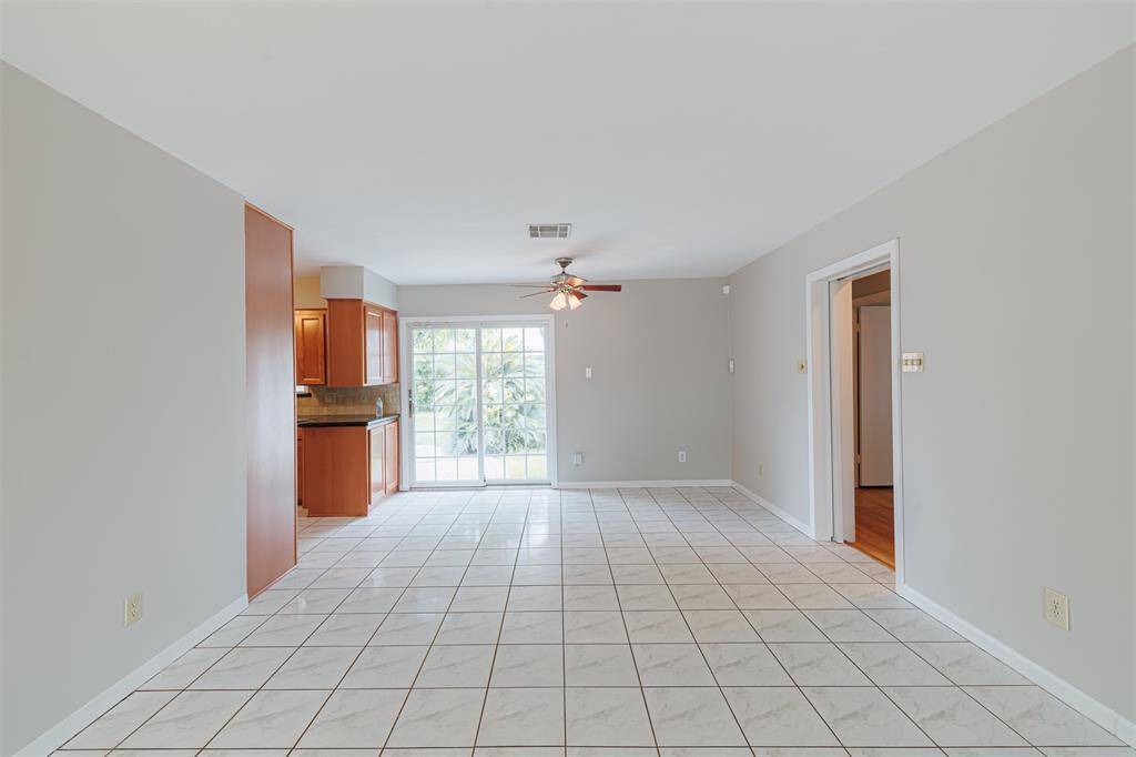 Bright, open living space with tiled floors, neutral walls, and a ceiling fan. It leads to a kitchen area with warm wood cabinetry and a large sliding glass door offering plenty of natural light and views of backyard.