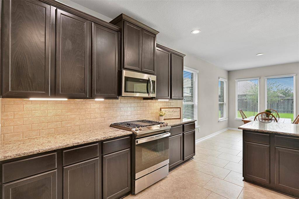 The kitchen features rich espresso cabinetry, under cabinet task lighting, granite countertops, a subway style backsplash, and stainless appliances including a gas range with a center griddle—all beautifully connected to the sunlit breakfast area with backyard views.