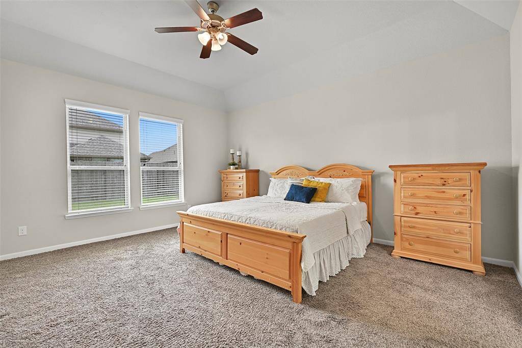 The spacious first-floor primary suite features plush carpeting, an elevated gambrel ceiling with a lighted ceiling fan, and abundant natural light from rear-facing windows overlooking the backyard.