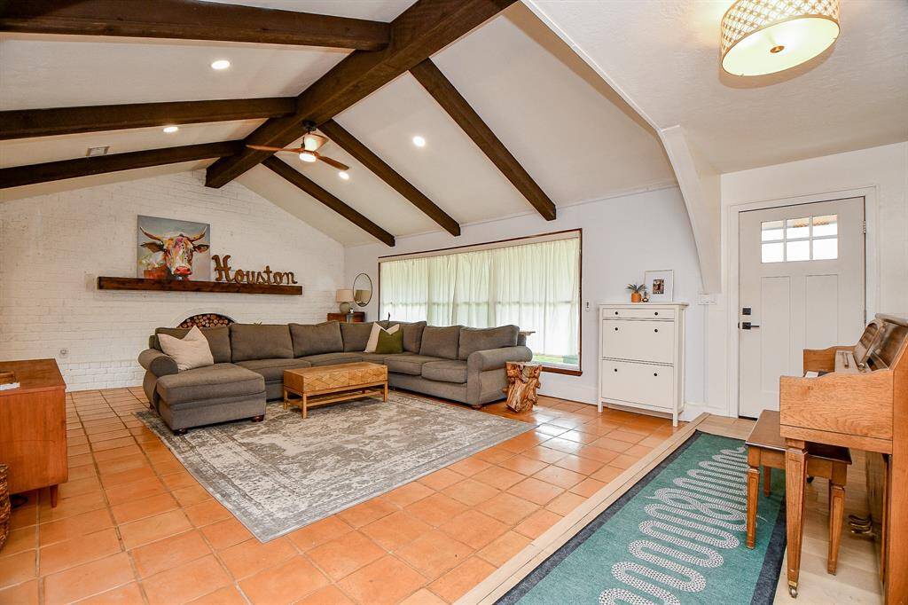 This inviting living room features a high ceiling with exposed wooden beams, a cozy sectional sofa, and warm terracotta tile flooring. A large window allows natural light, complementing the white brick accent wall and creating a bright, open space for relaxation.
