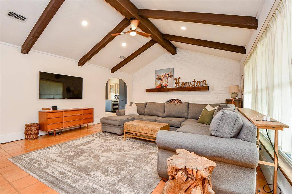 This spacious living room features a cozy sectional sofa, rustic wooden beams, and a bright atmosphere with large windows. It includes a mounted TV, stylish decor with a mix of modern and natural elements, and a decorative sign on a brick accent wall, creating a welcoming and comfortable space.