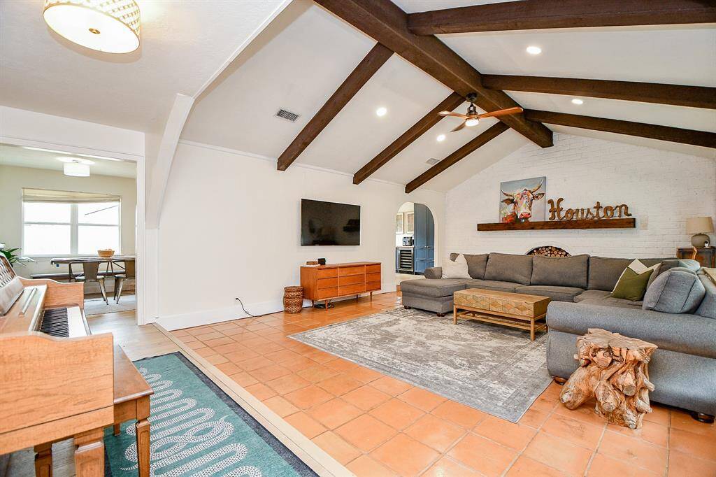 Spacious living room featuring exposed wooden beams, a cozy sectional sofa fits, and a rustic accent table. The area is well-lit with recessed lighting, and a large wall-mounted TV adds modern convenience. A piano and adjoining dining space enhance the open layout.