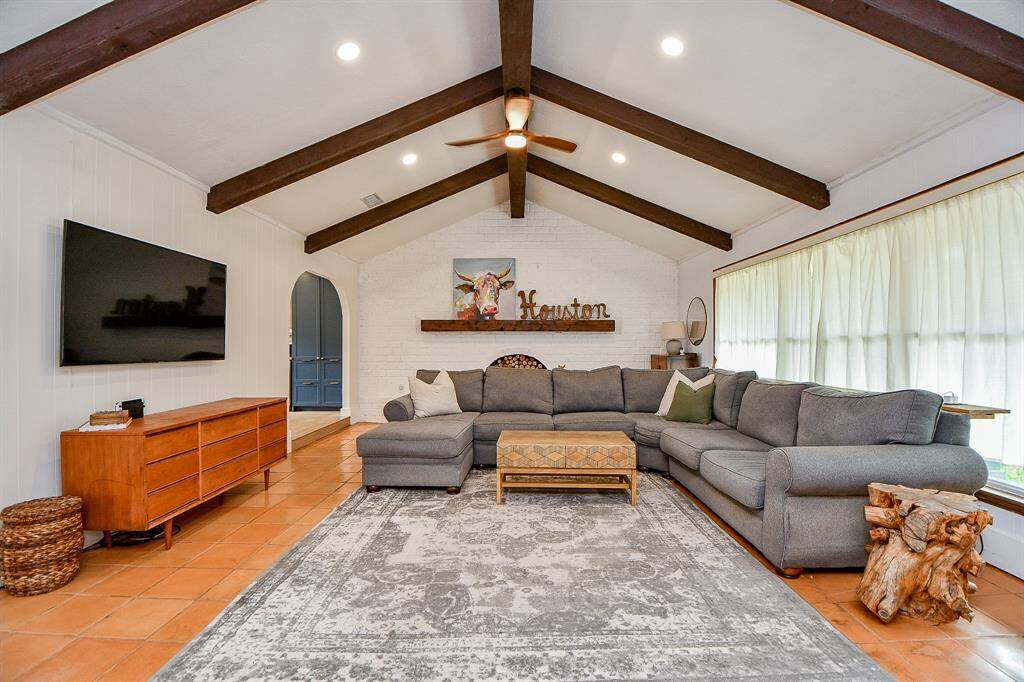 This spacious living room features vaulted ceilings with exposed wood beams, a cozy sectional sofa fits, and a large window allowing natural light to flood in. The room is a welcoming and comfortable space.