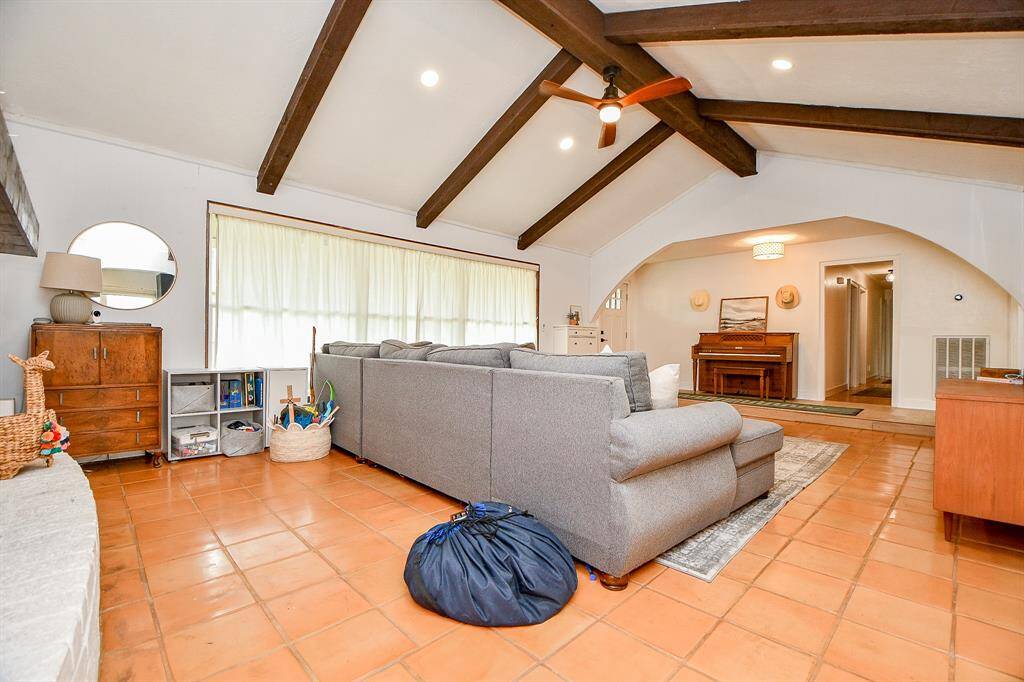 This inviting living room features a high ceiling with exposed wooden beams, terracotta tile flooring, and abundant natural light from a large window. A cozy gray sectional, warm wood accents, and a ceiling fan add comfort and style.