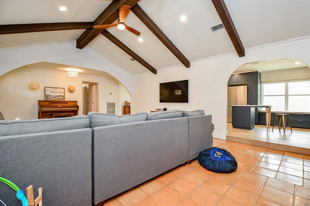 This inviting living space features a cozy sectional sofa, exposed wooden beams, and a ceiling fan. It opens into a kitchen with modern appliances and a breakfast bar. Warm terracotta tiles complement the open layout, creating a comfortable and stylish atmosphere.