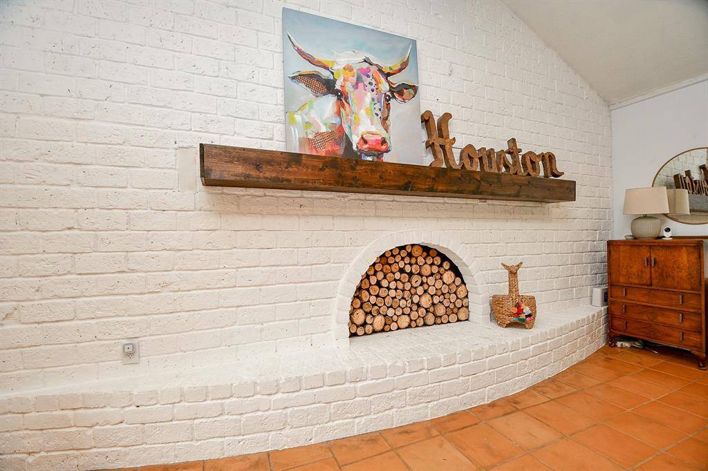 This photo showcases a cozy, rustic-style room with a white brick fireplace filled with logs. The wooden mantel features decorative accents and artwork. Terracotta floor tiles and a vintage wooden dresser add warmth and character to the space.