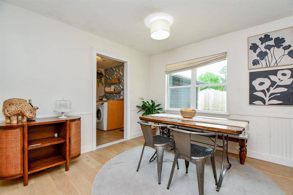 This bright dining area features a cozy wooden table with metal chairs, modern lighting, and large windows for natural light. A stylish sideboard and wall art add charm, with a view into a laundry room.