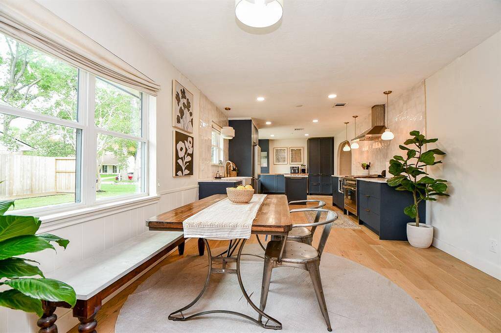 This photo showcases a bright, open-concept kitchen and dining area. It features a wooden dining table with metal chairs, large windows offering ample natural light, dark cabinetry, modern appliances, and light wood flooring, creating a warm and inviting space.