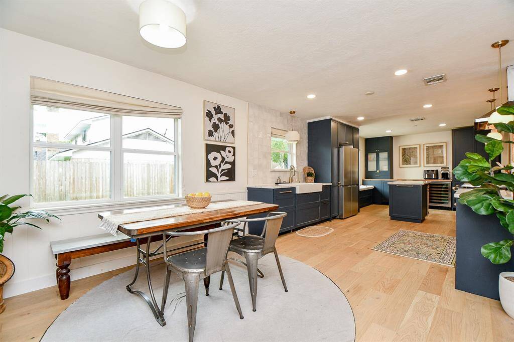 This photo showcases a modern, open-concept kitchen and dining area with light wood flooring. The kitchen features sleek dark cabinetry, stainless steel appliances, and a farmhouse sink. A bright dining space with large windows offers natural light, complemented by a stylish table and chairs. The area is accented with contemporary art and indoor plants, creating a welcoming atmosphere.