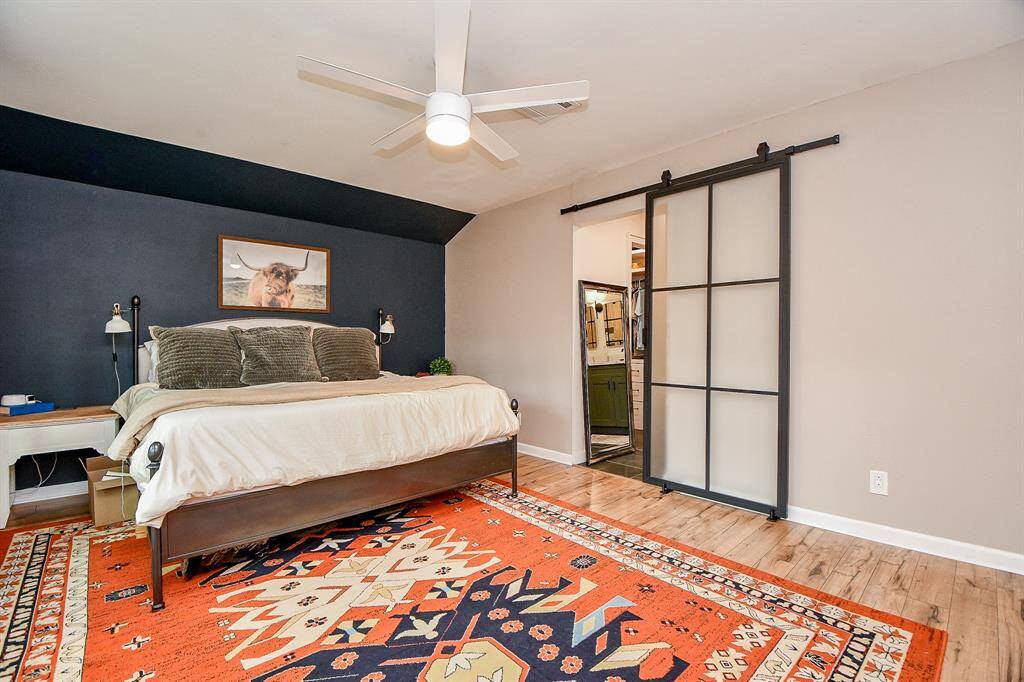This bedroom features a cozy, modern design with a large bed against a dark accent wall. It has a stylish ceiling fan, wood flooring, and a vibrant patterned rug. A sliding barn door leads to an adjacent space, adding a contemporary touch.