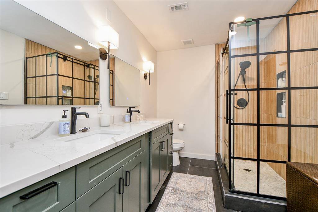 Modern bathroom featuring a double vanity with sleek black fixtures, a large mirror, and stylish lighting. The spacious shower has a glass enclosure with black grid accents and warm wood-like tiles.