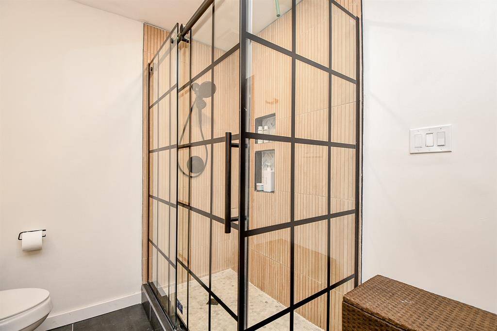 Modern bathroom with a sleek glass shower featuring black grid framing and wood-textured tile. The space is bright with a minimalist design, including a wall-mounted toilet and light switch panel.