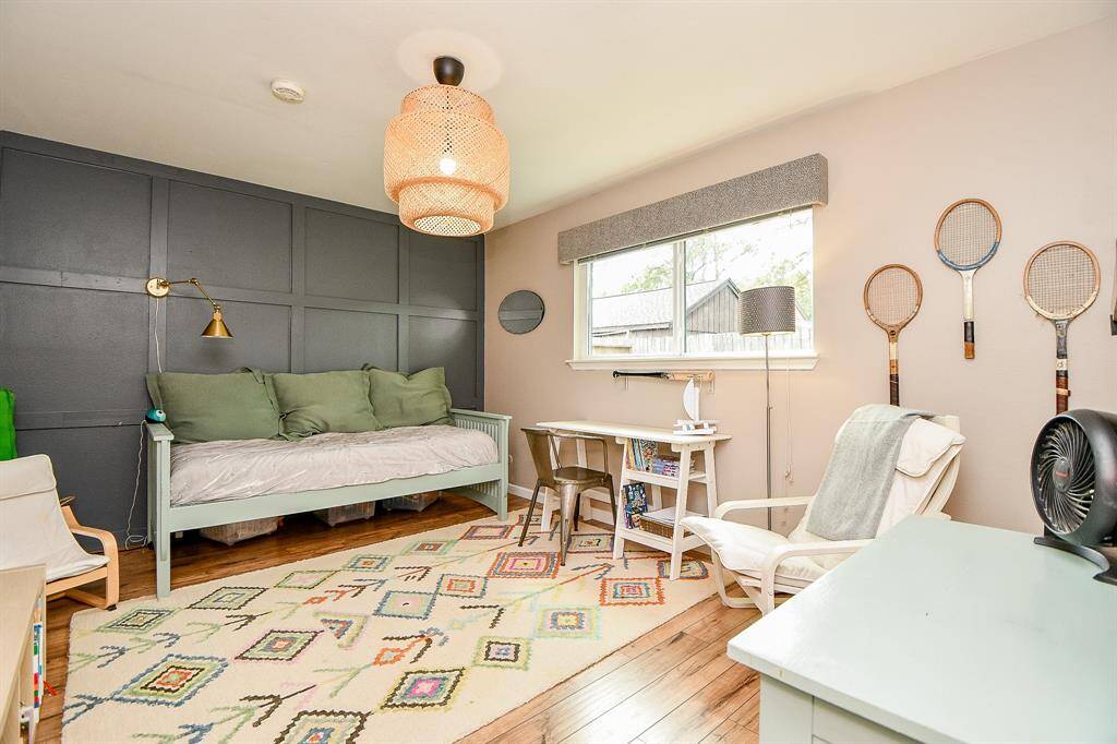 This cozy room features a dark accent wall with a daybed, stylish lighting, and a desk area by a large window. It has a warm wood floor, colorful area rug, and vintage tennis rackets as decor, creating a charming and inviting space.