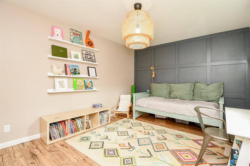 This cozy children's room features a stylish dark accent wall, a comfortable daybed with green cushions, and a playful, colorful rug. It includes wall-mounted shelves displaying books and decor, a low bookcase, and a modern woven light fixture, creating a welcoming and functional space.