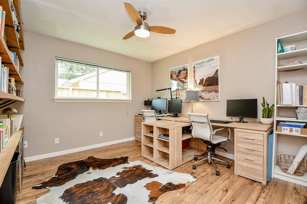 This home office features a spacious layout with a large window for natural light, wooden floors, and ample shelving. It includes a stylish ceiling fan and a dual desk setup, ideal for productivity.