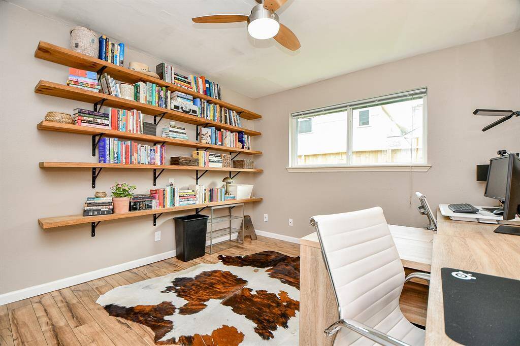 This bright home office features a large window, open shelving filled with books, a modern ceiling fan, and a stylish desk setup. The room has a warm wood floor and a cowhide rug, creating a cozy yet functional workspace.