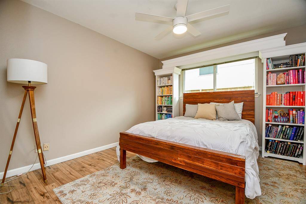 Cozy bedroom with warm wood flooring, a stylish wooden bed, and built-in bookshelves flanking a large window. The neutral walls and modern ceiling fan create a serene atmosphere, complemented by a decorative floor lamp and plush area rug.