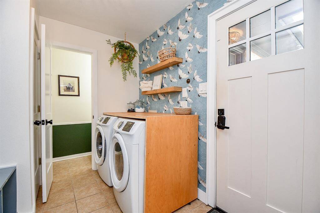 This photo showcases a bright and stylish laundry room featuring a modern washer and dryer set. The space is accentuated by a decorative wallpaper with a bird motif, open wooden shelving for storage, and a potted plant adding a touch of greenery. A door with glass panels provides natural light, enhancing the room's welcoming atmosphere.