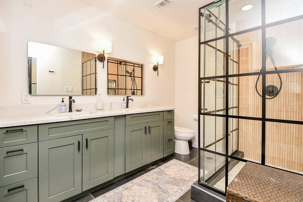 This bathroom features a modern, spacious design with green cabinetry, dual sinks, and sleek black fixtures. The large glass shower enclosure adds a touch of elegance, complemented by ample lighting and a neutral color palette.