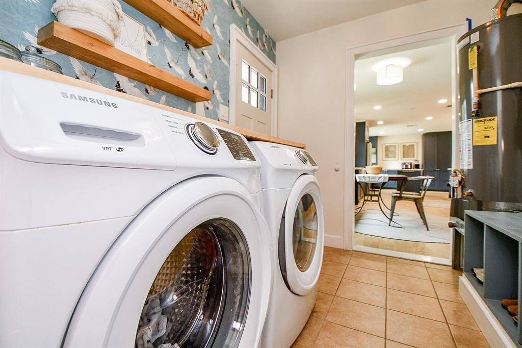 This photo showcases a well-lit laundry room featuring a modern washer and dryer set. The space is accented with stylish wallpaper and open wooden shelves, leading into a bright kitchen area.