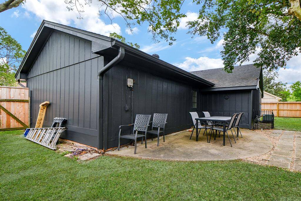 This photo shows a modern, dark-painted home exterior with a small patio area featuring outdoor seating. The yard is fenced and surrounded by greenery, providing privacy and a cozy outdoor space.
