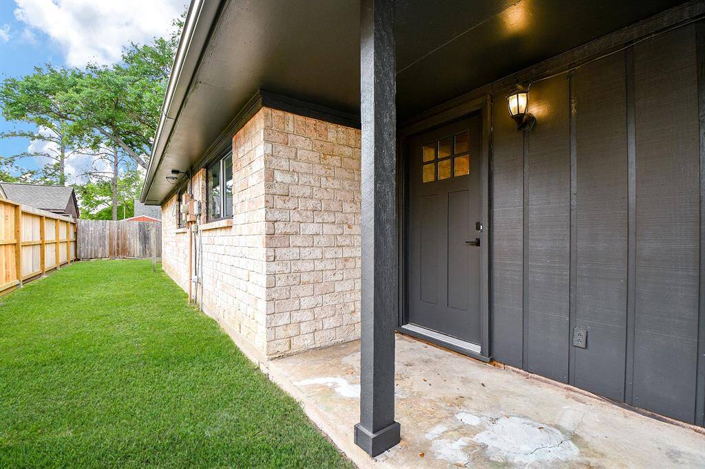 This photo shows a cozy entrance to a brick house with a modern black door and light fixture. There's a small, well-maintained front porch and a fenced yard with green grass, offering privacy and outdoor space.