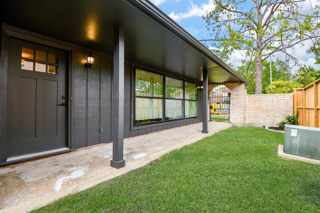 This photo showcases a charming covered porch with a modern dark exterior and large windows, leading to a private fenced yard with lush green grass and a brick wall, perfect for outdoor relaxation.