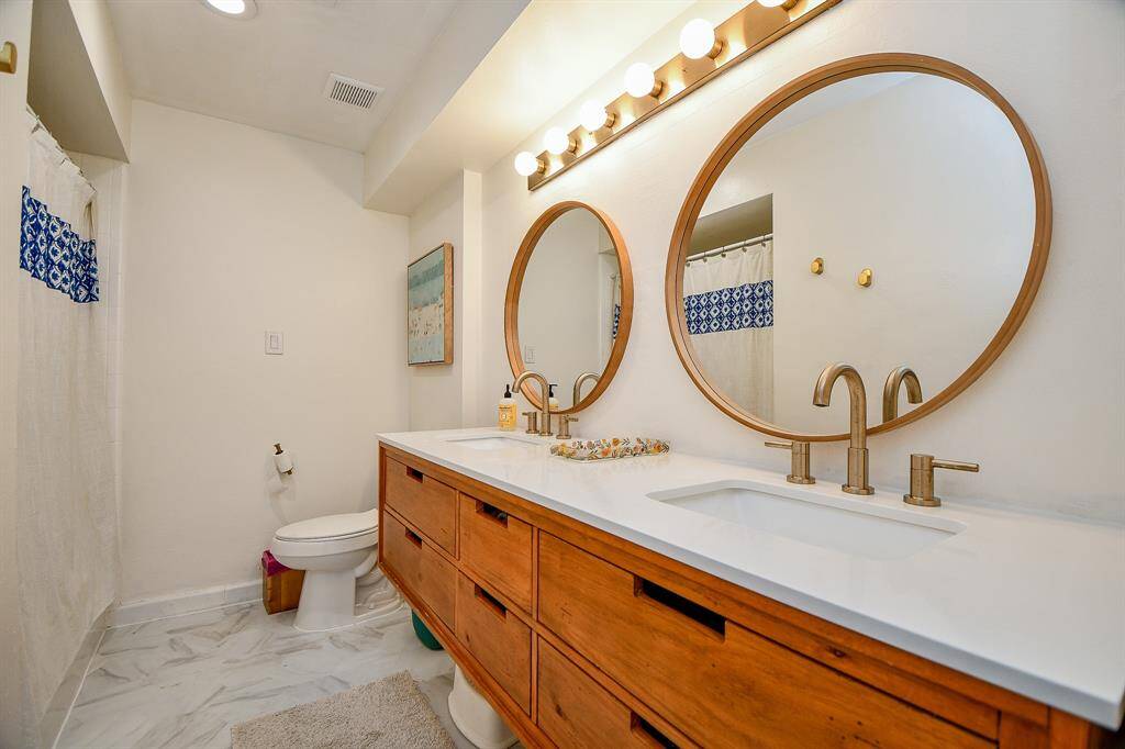 This bathroom features a modern design with dual round mirrors, a wooden vanity with double sinks, and brushed gold fixtures. The space is brightened by ample lighting and accented with a patterned shower curtain, creating a stylish and functional environment.