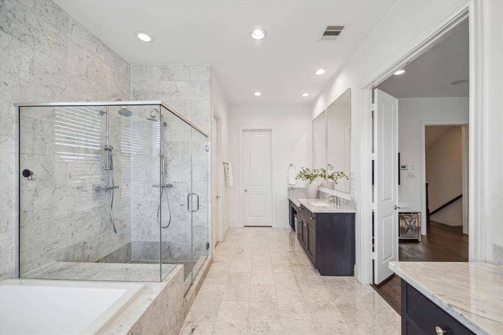 Unwind in the spa-inspired primary bath featuring an oversized walk-in shower with dual showerheads and sleek glass doors, dual vanities with generous counter space, and a deep soaking tub—all leading into a boutique-style walk-in closet complete with a custom-built island topped with marble and designed with additional storage.
