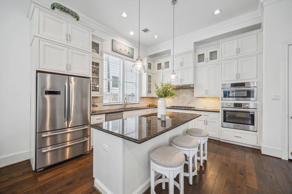 The island kitchen is the heart of the home, featuring Bosch stainless appliances, granite countertops, a gas cooktop, double oven, and abundant double stacked cabinetry with glass-front accents—plus a built-in beverage fridge tucked into the island for added convenience.