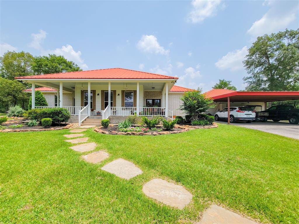 This charming home features a welcoming front porch with white railings, a vibrant red roof, and well-maintained landscaping. The spacious driveway leads to a covered carport, providing ample parking. The lush green lawn and stone path add to the home's inviting curb appeal.