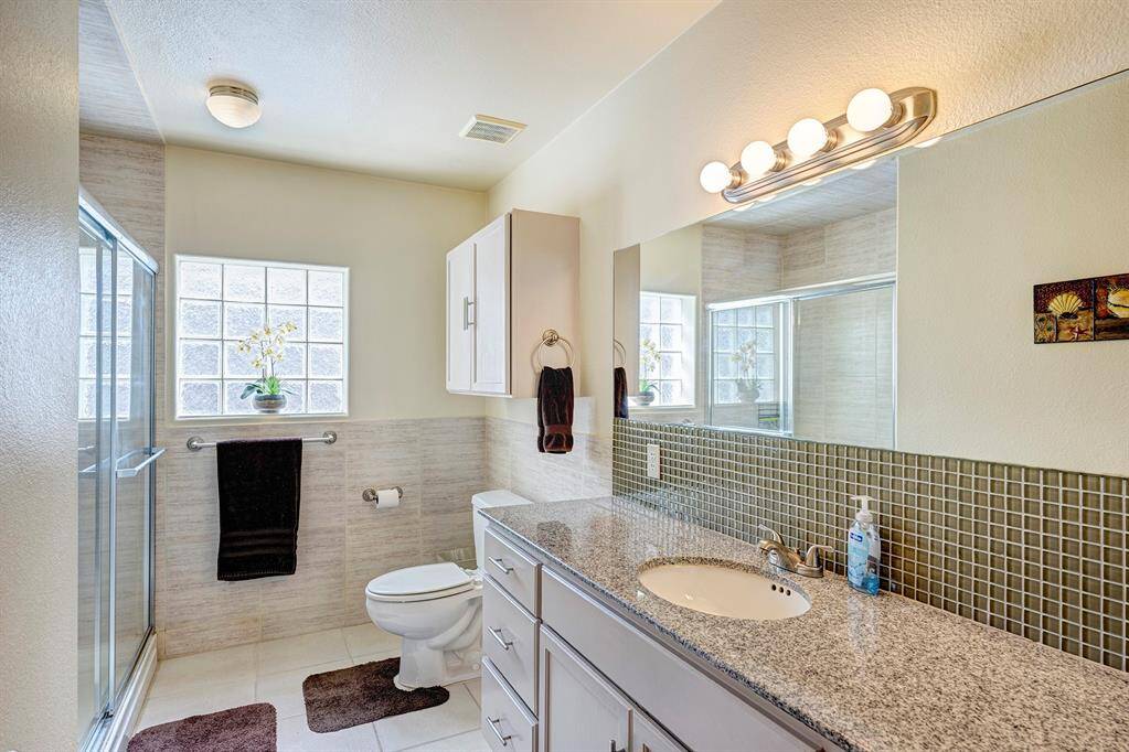 Bright, modern bathroom featuring a spacious glass-enclosed shower, granite countertop with a large mirror, and ample natural light through a glass block window.