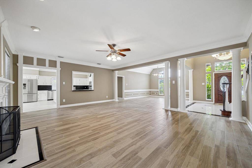 Bright and spacious open-concept living area with elegant flooring, ceiling fan, and large windows. It features a cozy fireplace, an inviting kitchen view, and a grand entrance with decorative columns and a stylish front door. Perfect for comfortable living and entertaining.