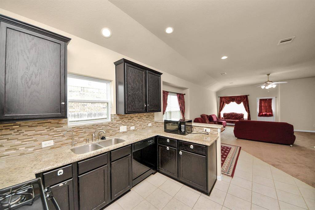 Spacious kitchen with dark cabinets, granite countertops, and a tiled backsplash opens to a cozy living area with red furnishings and large windows.