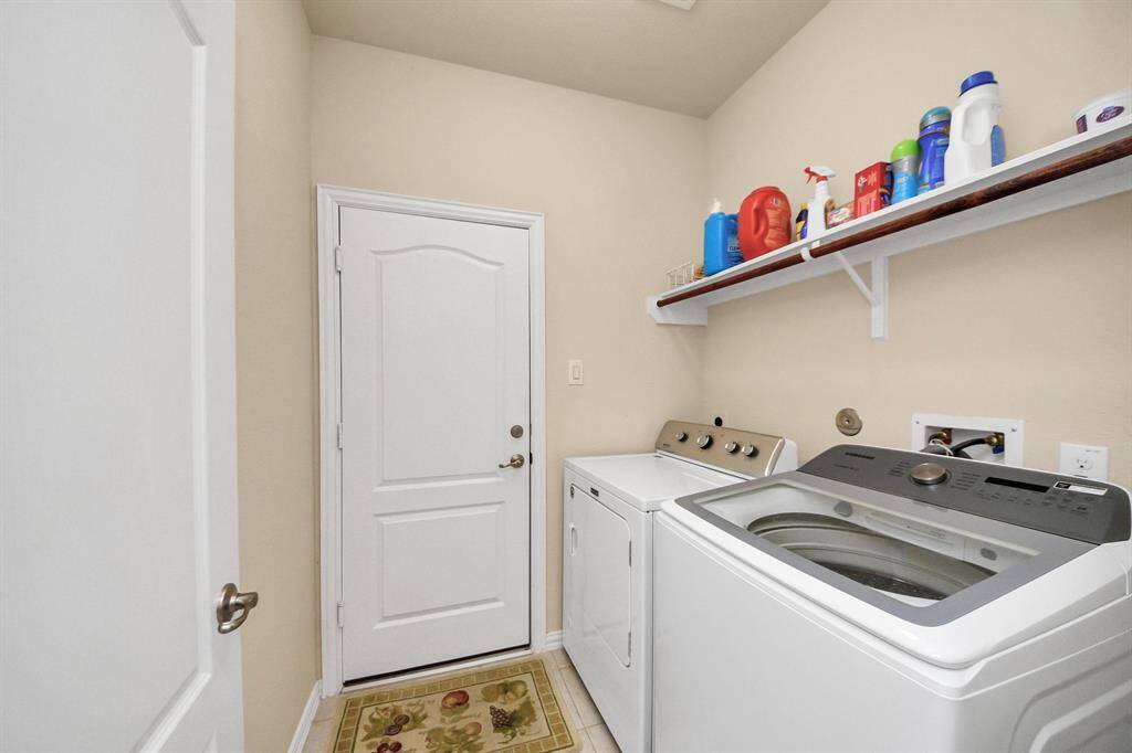 A compact laundry room with a washer and dryer, a shelf for storage, and a door leading to another area. The room is well-lit with light-color walls and tile flooring.