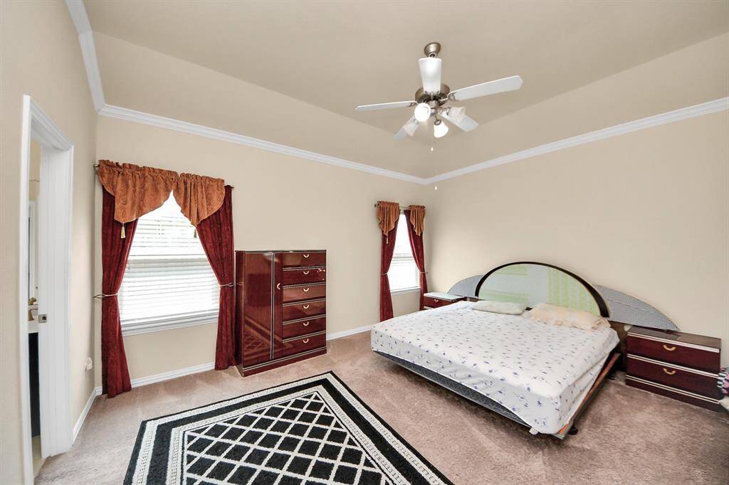 This primary bedroom features a spacious layout with neutral walls and carpet, a ceiling fan, and ample natural light from two windows. It includes elegant red drapes and classic wooden furniture, creating a cozy and inviting atmosphere.