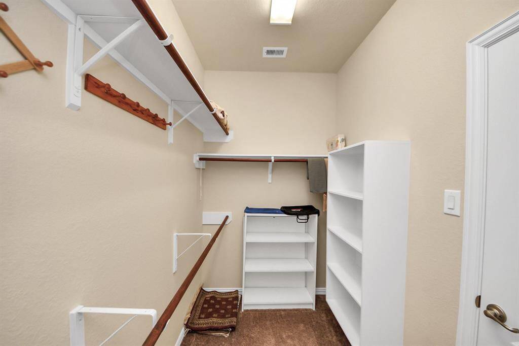 The primary walk-in closet with beige walls and carpeted flooring. It features multiple shelves and hanging rods for ample storage, with overhead lighting for visibility.
