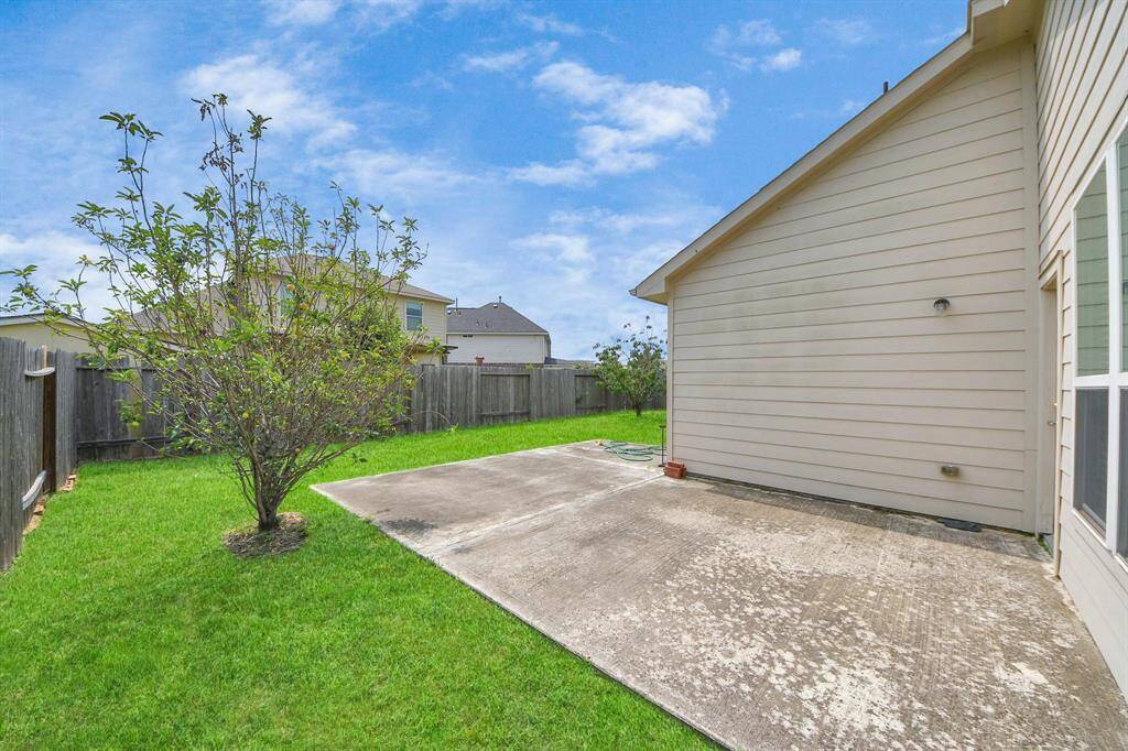 A backyard with a concrete patio adjacent to a house, a small tree, and a grassy area enclosed by a wooden fence. The space is open and well-maintained, ideal for outdoor activities.