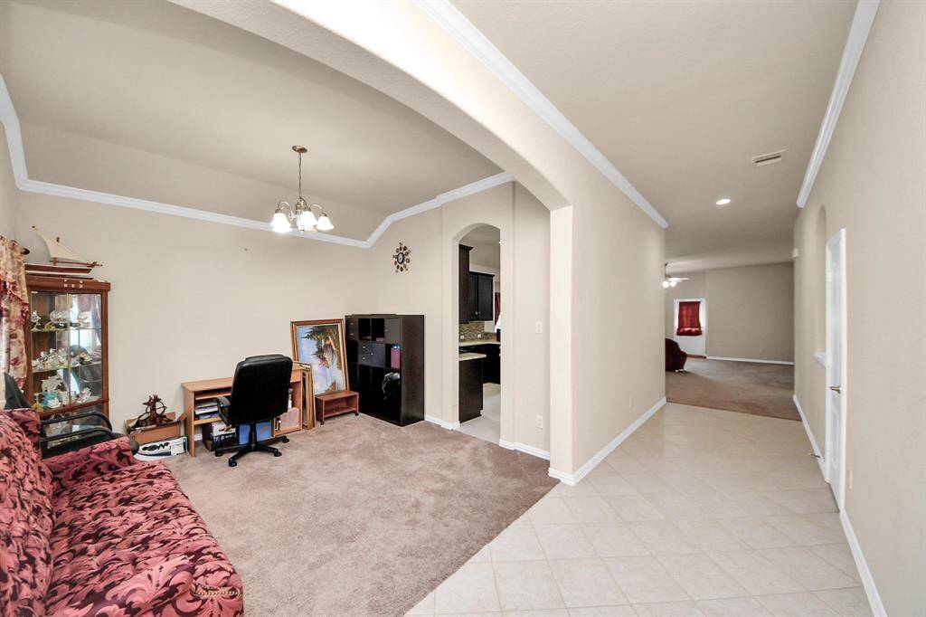 A tiled hallway with elegant archways leads to a cozy, carpeted room with a chandelier, desk, and bookshelf—bright, stylish, and full of charm.