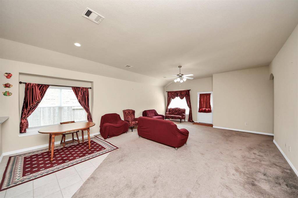 This spacious living room features neutral walls and carpet, with a ceiling fan and ample natural light from a large window adorned with red curtains. There are arched entryways and double doors leading to additional rooms, enhancing the open feel.