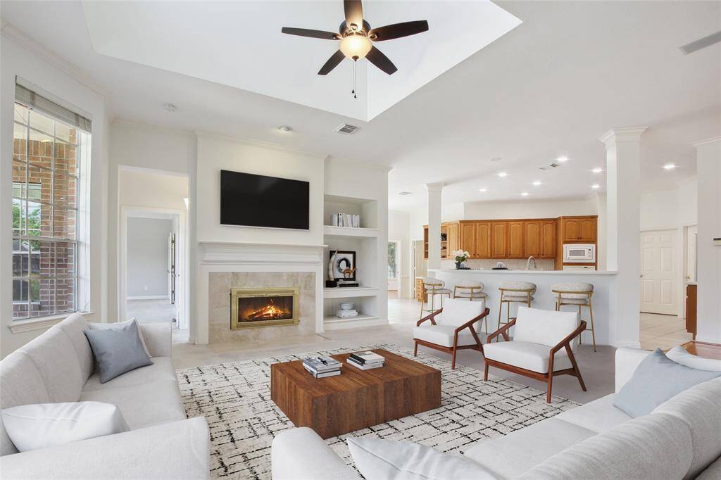 Spacious living area with a cozy fireplace and ceiling fan. Open layout connects to a modern kitchen with bar seating. Neutral tones and ample natural light create a welcoming atmosphere.