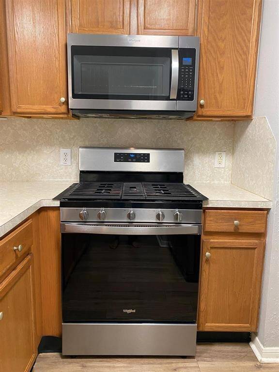 This photo showcases a modern kitchen with stainless steel appliances, including a fridge and microwave. It features a breakfast bar with a light countertop, wooden cabinetry, and recessed lighting. The space has a clean, contemporary feel with neutral walls and wood-look flooring.